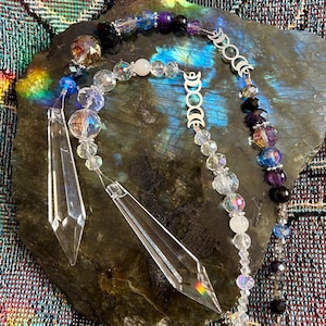 May include: A decorative crystal suncatcher with clear, iridescent, and purple beads, and two clear prism pendants. The suncatcher is displayed on a dark, textured stone surface, with a colorful, patterned background.
