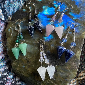 May include: A set of six pairs of dangle earrings featuring various gemstone pendants, including black, green, pink, blue, and white. Each pair has a silver chain with crystal beads.