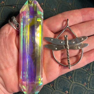 May include: A large, iridescent crystal point and a silver dragonfly pendant. The crystal is multi-faceted, reflecting a spectrum of colors. The dragonfly pendant features detailed wings and a circular frame.