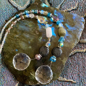 May include: A crystal suncatcher with two clear crystal prisms, black lava rock beads, and iridescent blue and pink beads hanging from a silver chain.