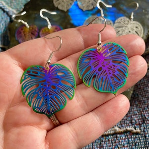 Pu&ograve; includere: Un paio di orecchini a forma di foglia di monstera arcobaleno iridescente. Gli orecchini sono in metallo e hanno un design delicato e intricato.