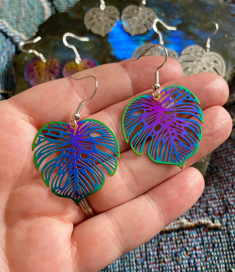 Pu&ograve; includere: Un paio di orecchini a forma di foglia di monstera arcobaleno iridescente. Gli orecchini sono in metallo e hanno un design delicato e intricato.