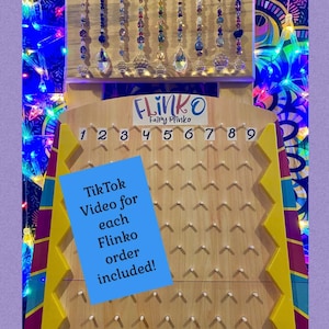 May include: A wooden Flinko game with a yellow and red frame. The game board has nine numbered slots and a series of pegs. A blue sticky note with the text "TikTok Video for each Flinko order included!" is attached to the game board. The game is decorated with colorful crystal suncatchers.