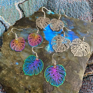 Pu&ograve; includere: Sei paia di orecchini a forma di foglia di monstera arcobaleno iridescenti. Gli orecchini sono in metallo e hanno un design delicato e intricato. Le foglie sono di un colore arcobaleno vibrante con una finitura metallica.