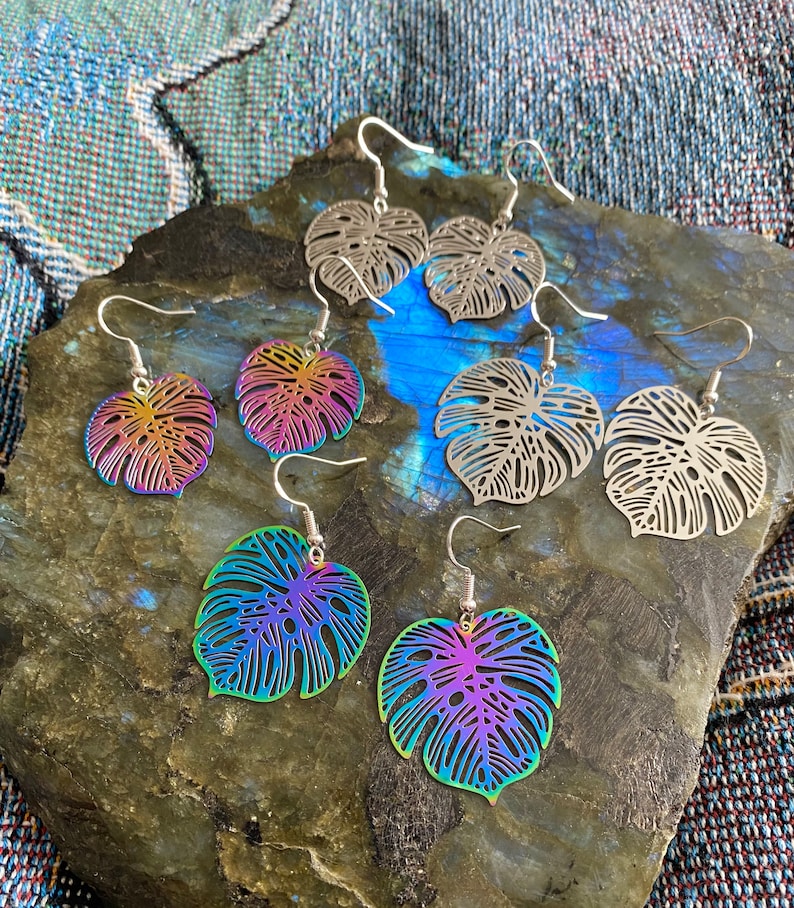 Pu&ograve; includere: Sei paia di orecchini a forma di foglia di monstera arcobaleno iridescenti. Gli orecchini sono in metallo e hanno un design delicato e intricato. Le foglie sono di un colore arcobaleno vibrante con una finitura metallica.