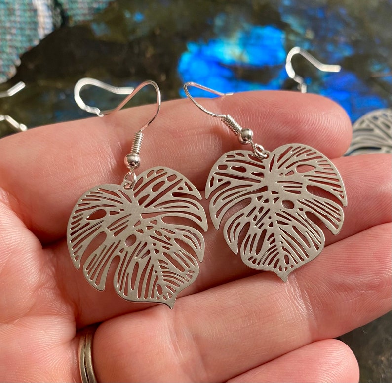 Pu&ograve; includere: Un paio di orecchini d'argento con un delicato design a taglio di una foglia di monstera. Gli orecchini sono di stile pendente con chiusura a gancio.