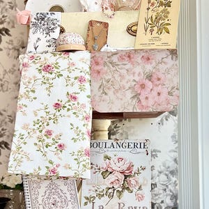 May include: A collection of vintage-style items, including floral-patterned fabrics, notebooks, and decorative cards. The color palette is soft, featuring pink, white, and gold tones. A framed picture and a teacup are also visible, creating a romantic aesthetic.