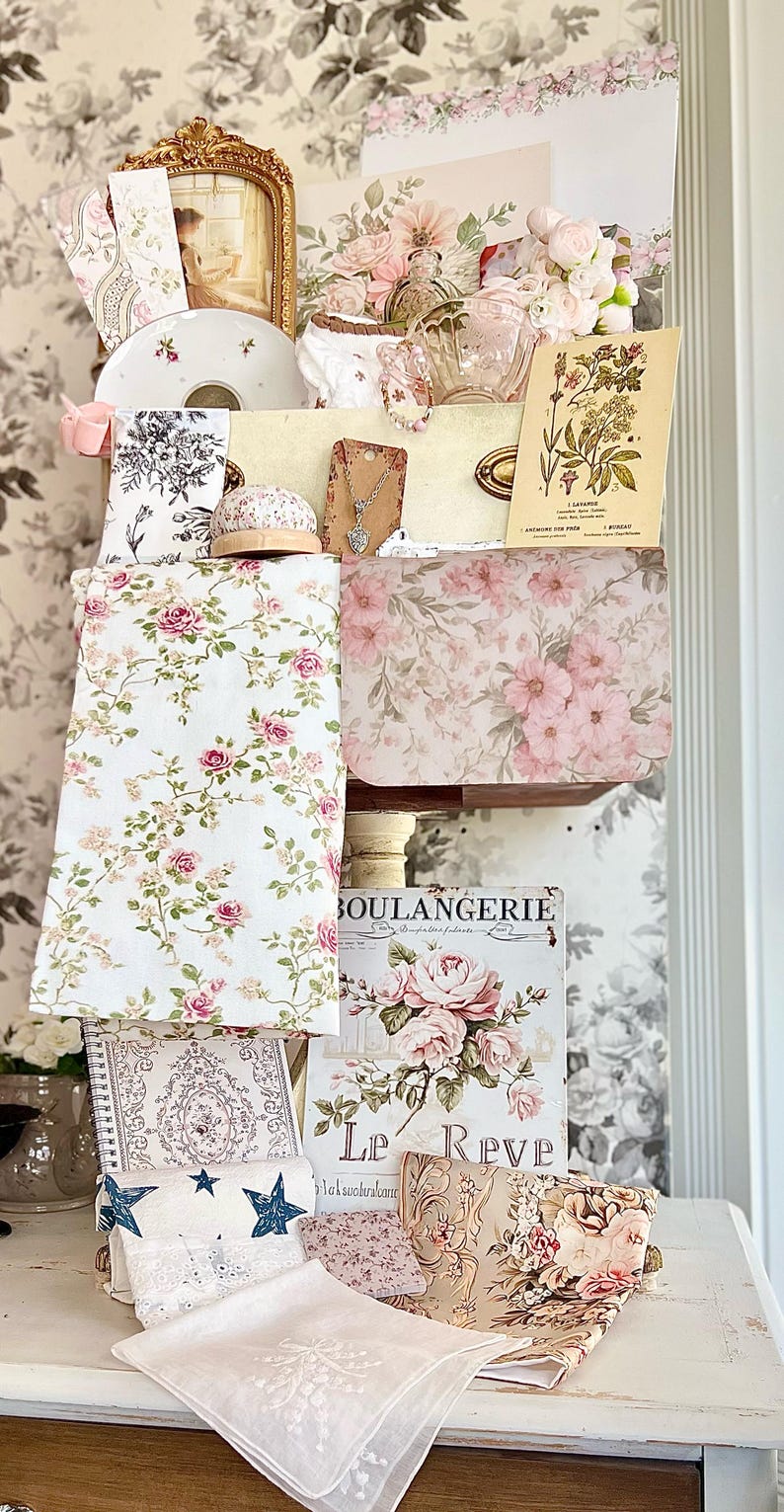 May include: A collection of vintage-style items, including floral-patterned fabrics, notebooks, and decorative cards. The color palette is soft, featuring pink, white, and gold tones. A framed picture and a teacup are also visible, creating a romantic aesthetic.