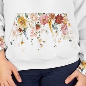 Op de afbeelding: Witte hoodie met een bloemenprint op de zak en de manchetten. De bloemen zijn in tinten roze, geel en rood.