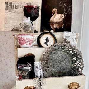 May include: A multi-tiered shelf displays vintage decor. Items include a red glass goblet, a swan print, a teacup with a red and white pattern, a silhouette in a black frame, and a decorative metal plate. The shelf has brass-toned drawer pulls.