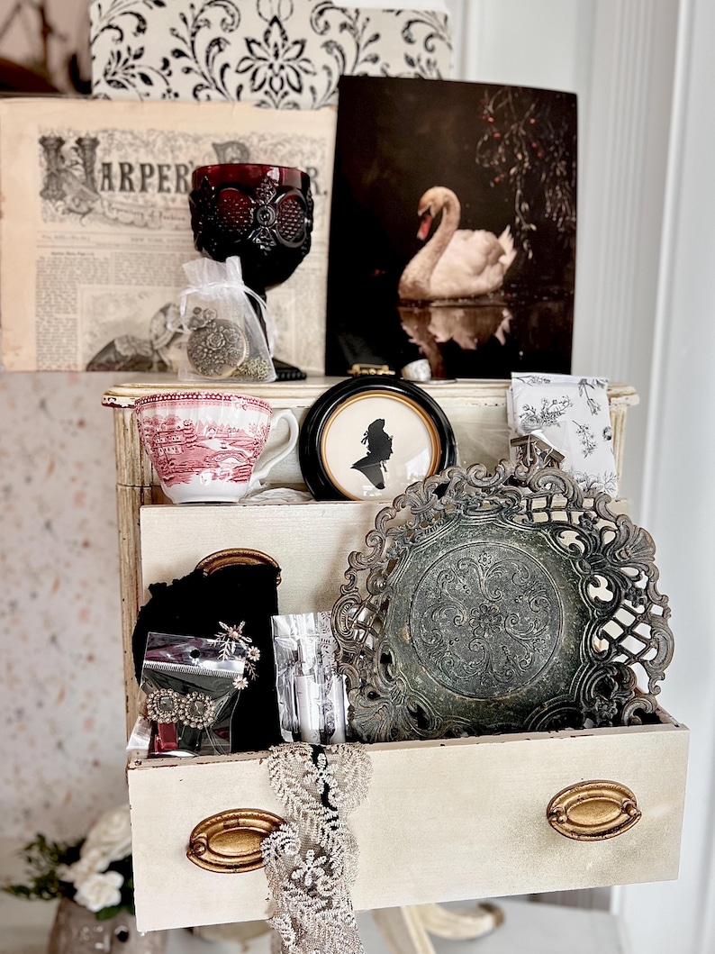 May include: A multi-tiered shelf displays vintage decor. Items include a red glass goblet, a swan print, a teacup with a red and white pattern, a silhouette in a black frame, and a decorative metal plate. The shelf has brass-toned drawer pulls.
