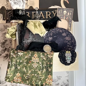 May include: A curated collection of vintage-inspired decor items. Includes a black raven figurine, floral patterned fabric, a black framed picture, and a decorative owl brooch. The items are arranged on a shelf with a floral wallpaper background.