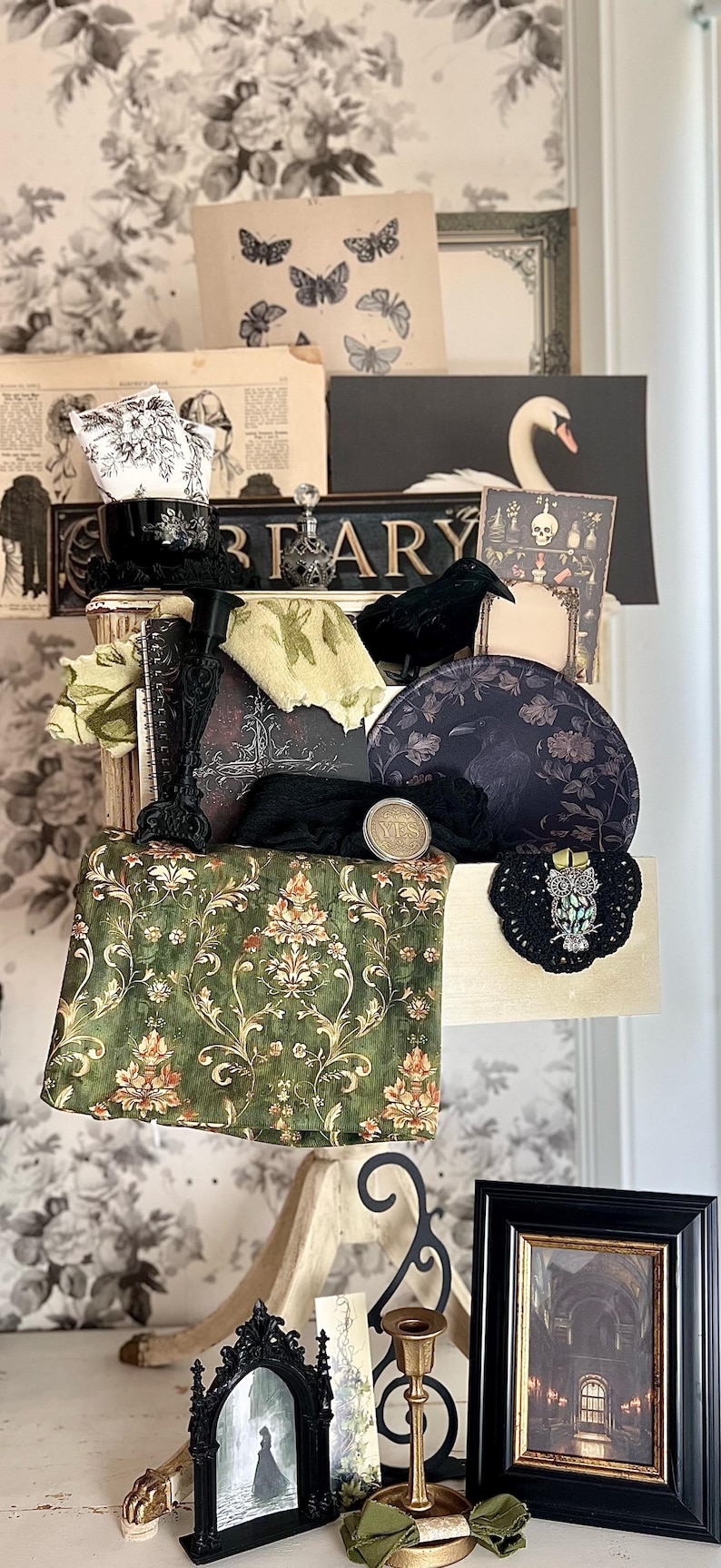 May include: A curated collection of vintage-inspired decor items. Includes a black raven figurine, floral patterned fabric, a black framed picture, and a decorative owl brooch. The items are arranged on a shelf with a floral wallpaper background.