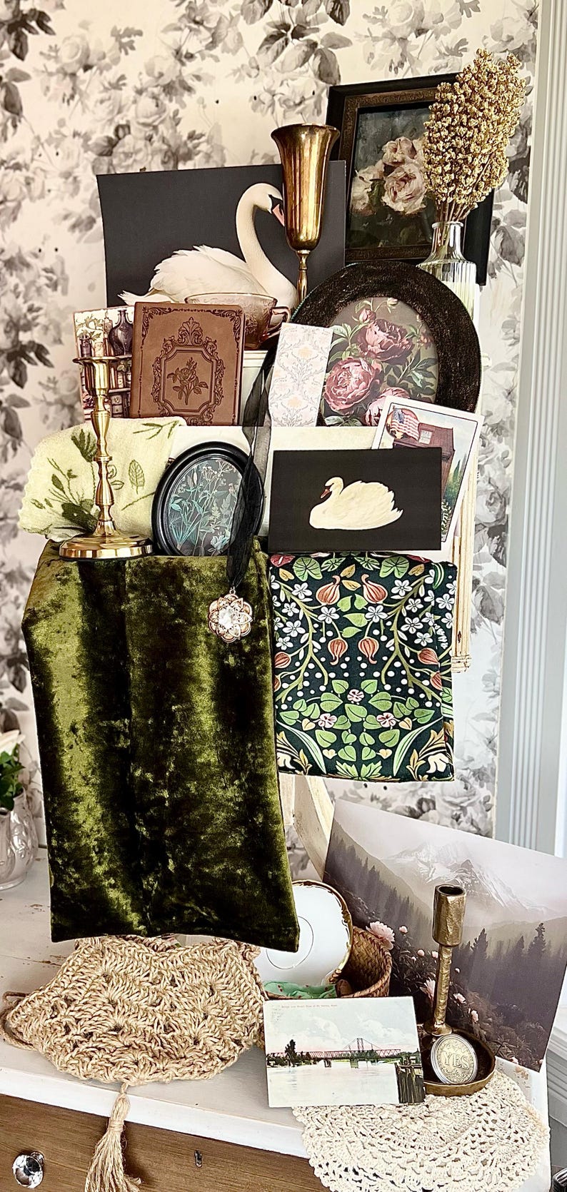 May include: An assortment of vintage-inspired decor items, including a swan illustration, framed floral prints, a velvet green fabric, and a gold candlestick. A woven bag and a postcard are also visible.