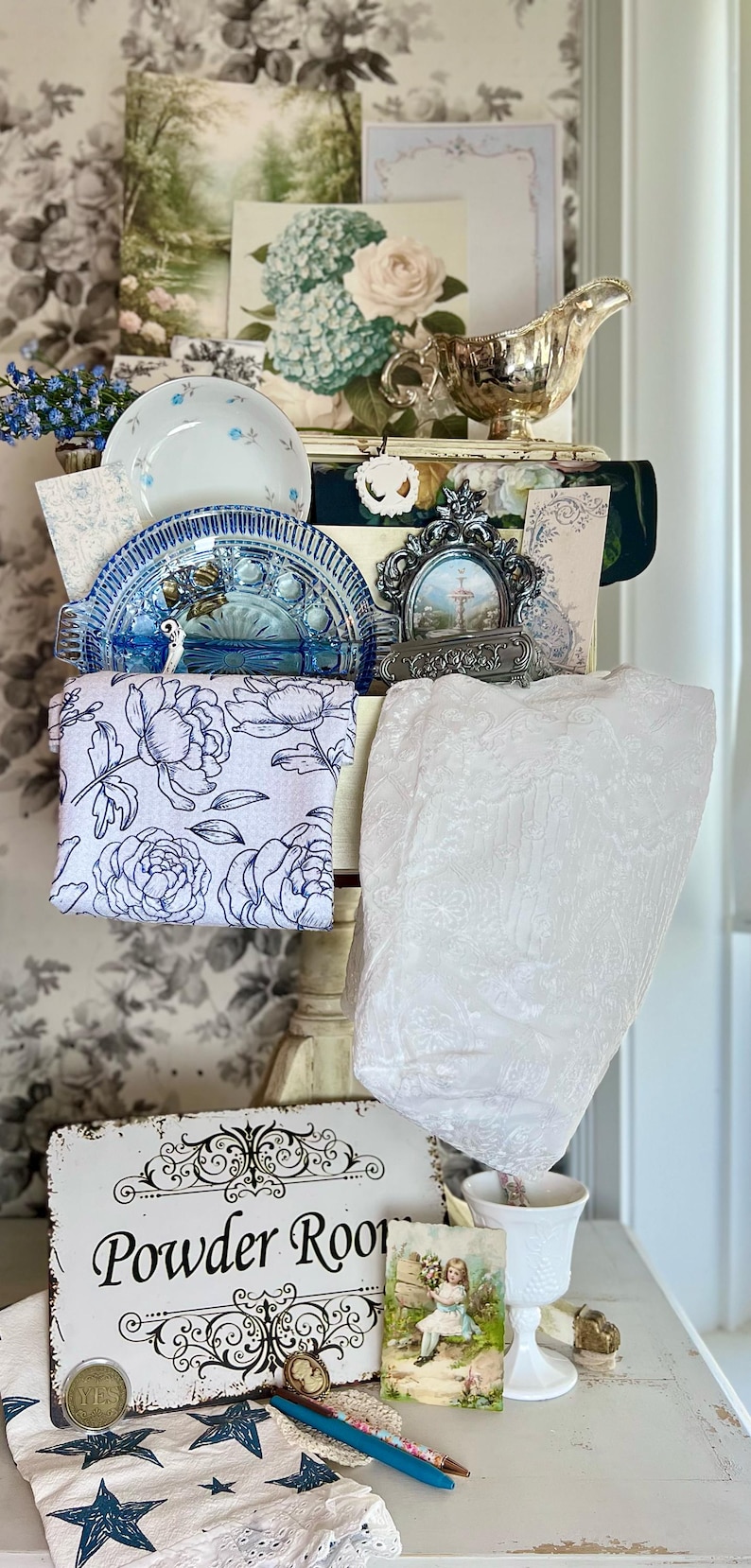May include: A display of vintage decor items, including a silver gravy boat, blue and white china, floral artwork, and a sign that reads "Powder Room." A white lace fabric and a star-patterned cloth are also visible.