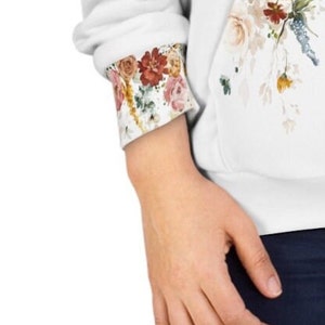 Op de afbeelding: Witte hoodie met een bloemenprint op de voorkant en de manchetten. De bloemenprint heeft roze, rode en gele bloemen.