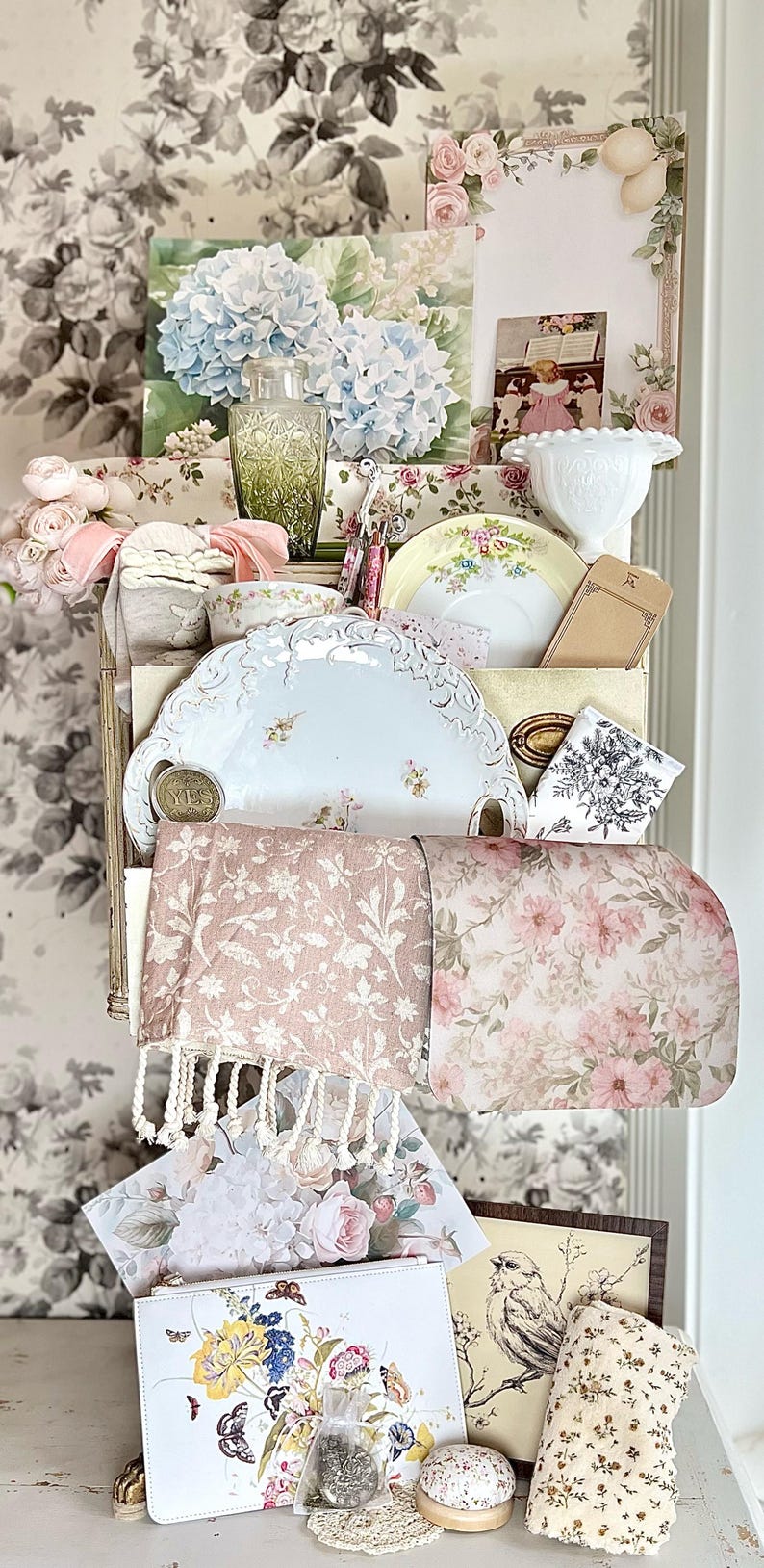 May include: A display of vintage-style items, including floral-patterned plates, a green glass bottle, and a clutch with a butterfly design. The backdrop features a floral wallpaper, creating a cohesive aesthetic.