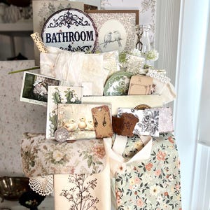 May include: A collection of vintage-style items, including floral-patterned fabric, decorative cards, and a metal sign that reads "Bathroom." The items are arranged on a white surface, creating a charming, nostalgic aesthetic. The color palette includes soft pastels and muted tones.