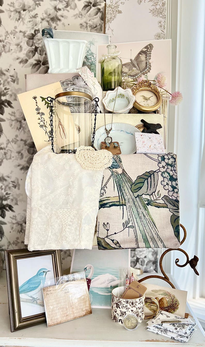 May include: A collection of vintage-style decorative items, including a framed blue bird print, a white lace cloth, a green glass bottle, and a floral patterned mug. Other items include a small gold framed picture, a decorative plate, and a butterfly print.