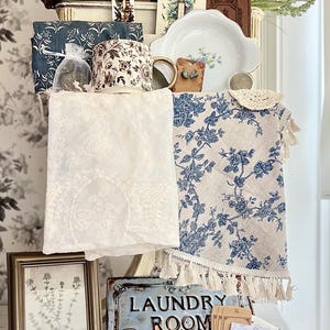 May include: A collection of vintage-style home decor items, including a woven basket, floral-patterned fabric, a framed botanical print, and a metal "LAUNDRY ROOM" sign. The color palette includes white, blue, brown, and gold tones, creating a rustic and charming aesthetic.