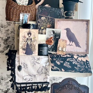 May include: A collection of vintage-inspired items, including framed art, a woven basket, a black crow figurine, and a candle. The items are arranged on a white surface against a floral wallpaper backdrop. The color palette includes black, white, and touches of gold.