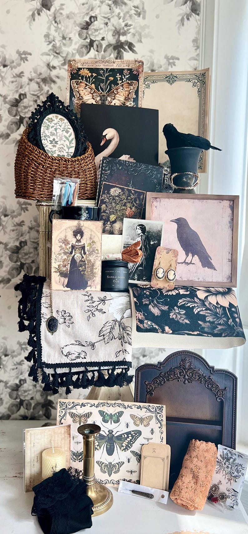 May include: A collection of vintage-inspired items, including framed art, a woven basket, a black crow figurine, and a candle. The items are arranged on a white surface against a floral wallpaper backdrop. The color palette includes black, white, and touches of gold.