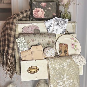 May include: A display of vintage-style items on a white stand. Includes a framed rose print, floral-patterned wrapping paper, a decorative plate with pink flowers, and a collection of small envelopes. A brown and cream plaid throw is draped over the display.