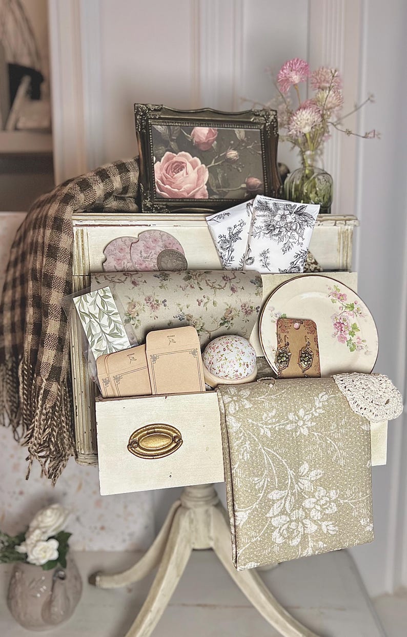 May include: A display of vintage-style items on a white stand. Includes a framed rose print, floral-patterned wrapping paper, a decorative plate with pink flowers, and a collection of small envelopes. A brown and cream plaid throw is draped over the display.