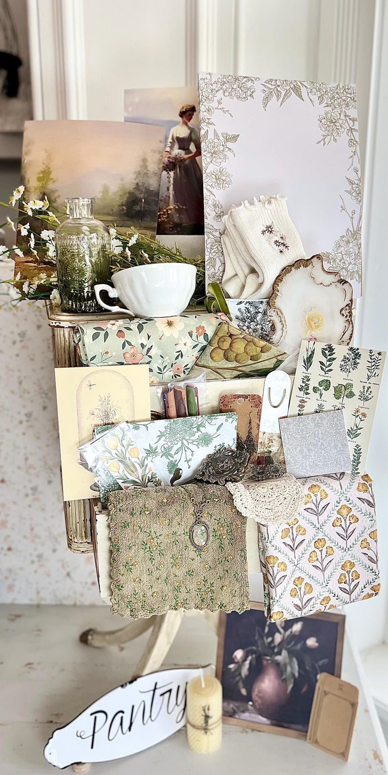 May include: A collection of vintage-style items, including a teacup, floral-patterned fabric, postcards, and a small painting. The items are arranged in a decorative, antique-style storage unit. A sign that says "Pantry" is visible.