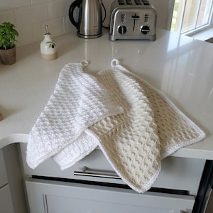 May include: Two rectangular kitchen towels featuring a textured waffle-knit pattern. One towel is solid white, and the other is a light beige colour. Each towel includes a small white loop at the top corner for hanging, with finished edges.