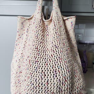 May include: Cream-colored knitted tote bag with a mesh design. The bag has colorful flecks of red, blue, and purple throughout the knit. The bag has two handles and is hanging from a metal hook.