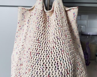 May include: Cream-colored knitted tote bag with a mesh design. The bag has colorful flecks of red, blue, and purple throughout the knit. The bag has two handles and is hanging from a metal hook.