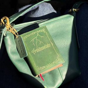 Può includere: Una borsa in pelle verde con un piccolo libro che recita "Frankenstein" in lettere dorate. Il libro ha un segnalibro rosso. Un'etichetta in pelle verde pende dalla catena dorata della borsa.