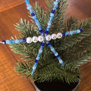 May include: A blue and white beaded snowflake ornament with the name "JAYSON" spelled out in white beads with gold lettering.