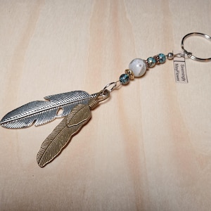 May include: A silver and gold feather keychain with turquoise and white beads and a white tag that reads "Seemingly MayFlower".