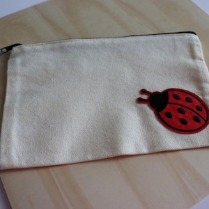 May include: Cream-colored canvas pouch with a black zipper and a red ladybug applique. The ladybug has black spots and a black head. The pouch is rectangular and appears to be made of a sturdy fabric.
