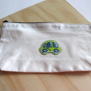 May include: A cream-colored pencil case with a black zipper and a small fabric loop. The case features an embroidered green car with blue accents. The case is rectangular and rests on a light wood surface.
