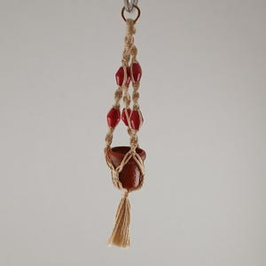 Op de afbeelding: Een handgemaakte macramé plantenhouder met een kleine terracotta pot. Het beige macramé heeft rode kralen en een kwast. De pot hangt aan een metalen ring, perfect voor kleine planten.
