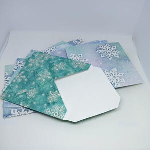 May include: A stack of square envelopes featuring a watercolor-style design in shades of teal, blue, and purple. The envelopes are decorated with white snowflake patterns of varying sizes. One envelope is open, revealing a plain white interior.