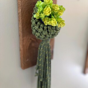 Könnte beinhalten: Ein handgefertigter Makramee-Pflanzenaufhänger in Olivgrün mit Goldpunkten, der einen Strauß künstlicher gelb-grüner Blumen hält. Der Aufhänger hat eine lange Quaste und ist auf einer Holzplanke montiert.