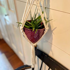 Pode incluir: Um suporte de plantas macramé branco com um vaso de cerâmica bordô que contém uma planta de aloe vera verde. O suporte de plantas está suspenso contra uma parede branca, com uma cadeira preta em primeiro plano.