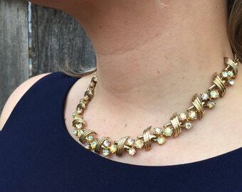 May include: A gold necklace with iridescent stones set in a criss-cross pattern. The necklace is worn by a person with a dark blue top.