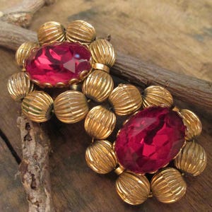 May include: A pair of gold tone earrings with red rhinestone accents. The earrings are made of round, textured metal beads.