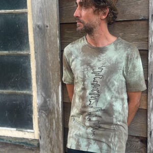 May include: An olive green t-shirt with a tie-dye pattern and a black line drawing down the front. The model wears black trousers and a brown tweed hat. The background features a wooden wall and a window.