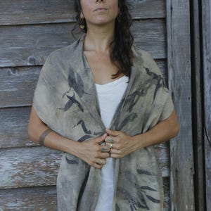 Puede incluir: Una bufanda beige con un estampado de hojas gris oscuro. La bufanda está drapeada sobre los hombros y tiene un borde con flecos. La persona de la imagen lleva una camiseta blanca.