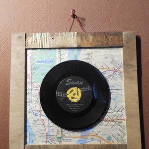 May include: A framed vintage record with a black vinyl record and a yellow label. The label has the text "Swan" and "She Loves You" by "The Beatles". The record is mounted on a map of Manhattan, New York City.
