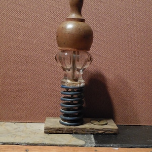 May include: A wooden bottle stopper with a clear glass base and a blue spring. The stopper is on a wooden base with a metal screw.