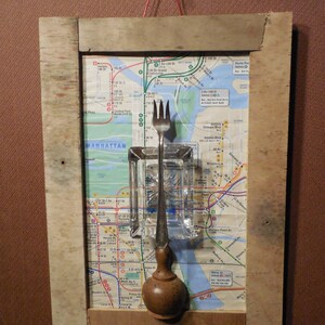 May include: A vintage fork and a glass bottle stopper are displayed on a map of the New York City subway system. The map is mounted on a wooden frame. The fork is silver and the bottle stopper is brown.