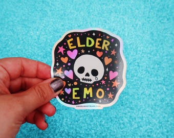 May include: A holographic sticker with a black background and the words "ELDER EMO" in yellow and purple. The sticker features a white skull with a teardrop, surrounded by hearts and stars in pink, orange, and yellow. The website CECILIAHOTZLER.COM is printed at the bottom.