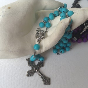 Puede incluir: Un rosario azul turquesa con un crucifijo de plata y un dije de plata con una imagen de Jes&uacute;s. El rosario est&aacute; sobre una superficie blanca.